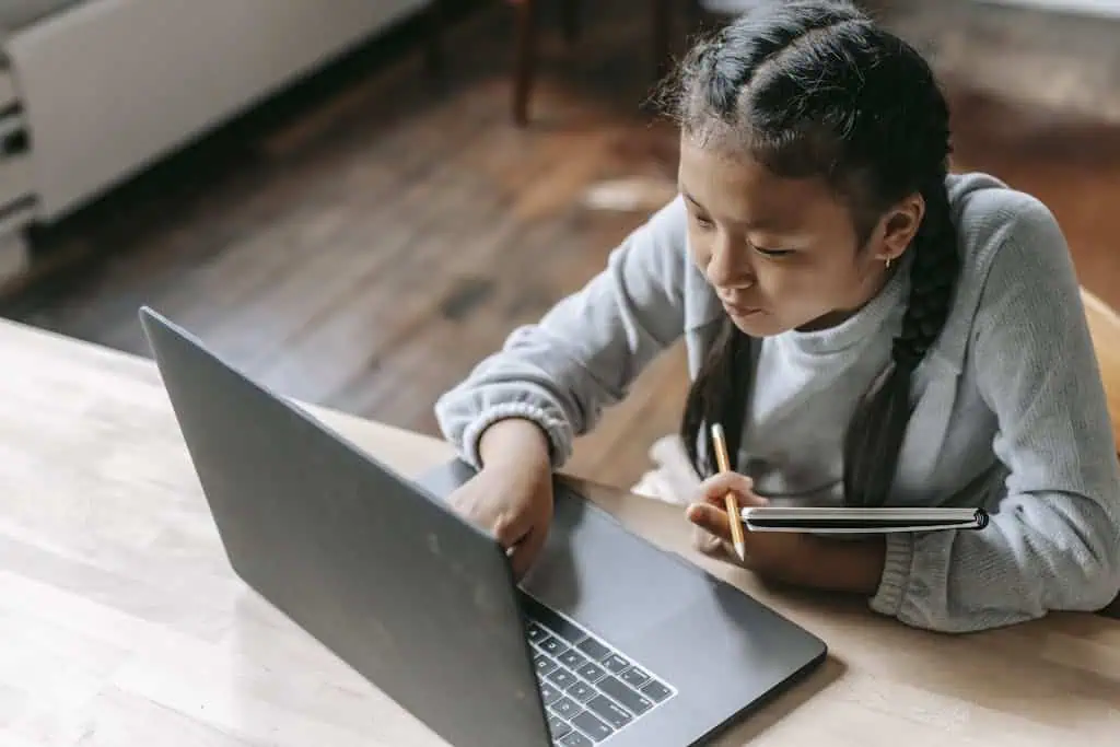 From above of concentrated Asian schoolgirl with pigtails sitting at wooden table with copybook and pencil and browsing laptop for school task
