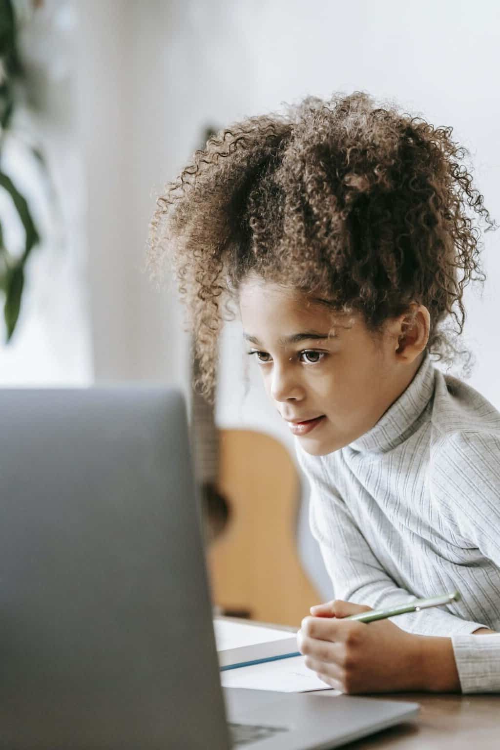Positive African American girl with curly hair taking notes on paper while looking at screen of netbook during studies at home with guitar on blurred background
